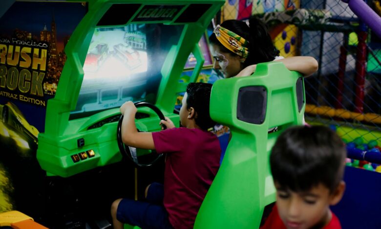 Unrecognizable child having fun with bright car racing simulator while turning steering wheel near serious ethnic woman and funny brother in shopping center near playground