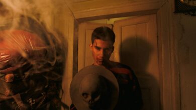 A man in a dimly lit room holds a hat adorned with a skull, surrounded by smoke.