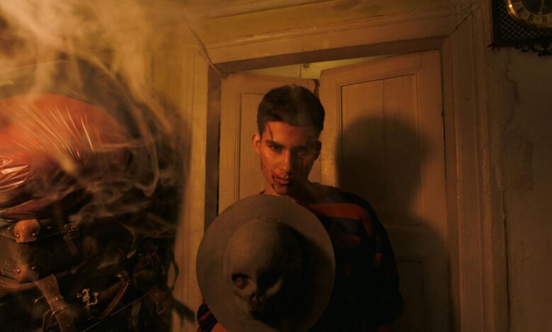 A man in a dimly lit room holds a hat adorned with a skull, surrounded by smoke.