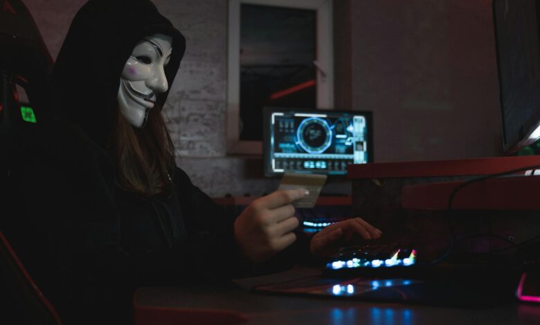 Masked hacker with credit card at computer, symbolizing cybercrime and anonymity.