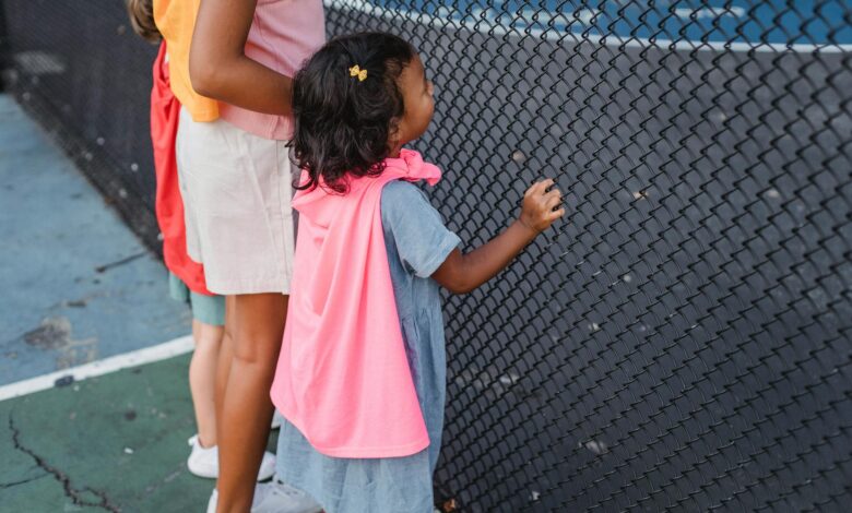 Kids wearing colorful capes watch eagerly through a fence, capturing the joy of outdoor play.