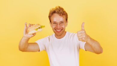 Smiling man in white shirt holding a burger against bright yellow background. Thumbs up gesture adds positive vibe.