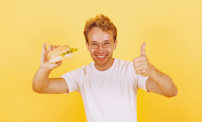 Smiling man in white shirt holding a burger against bright yellow background. Thumbs up gesture adds positive vibe.