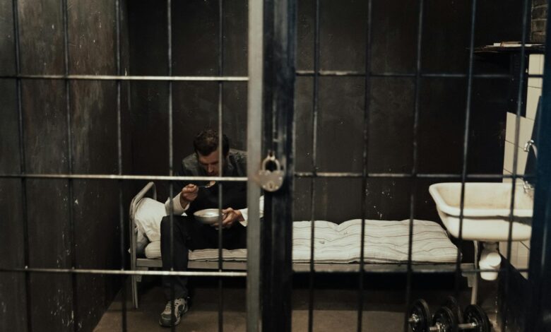A prisoner sits alone in a dark cell, eating with focus in a moody environment.