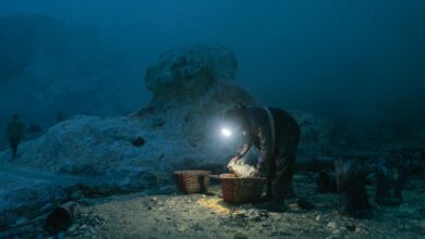 A solitary miner with headlamp gathers sulfur in baskets at a quarry during night.