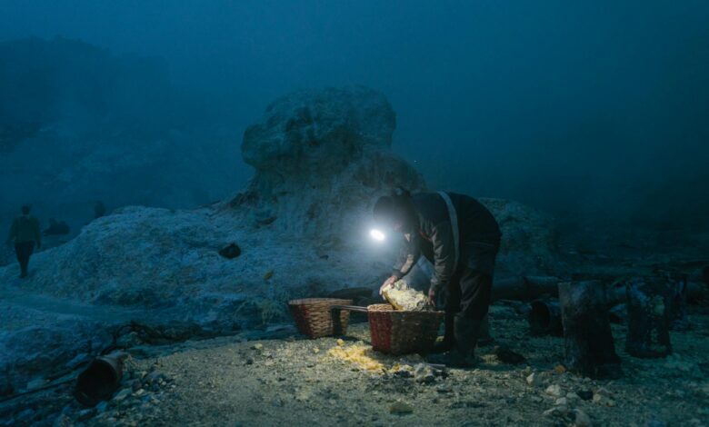 A solitary miner with headlamp gathers sulfur in baskets at a quarry during night.