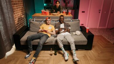 Three friends enjoying a gaming night in a cozy living room with colorful lights and drinks.