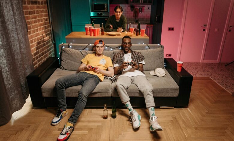 Three friends enjoying a gaming night in a cozy living room with colorful lights and drinks.