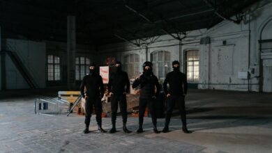 Four individuals in balaclavas pose in an empty warehouse setting, exuding a sense of secrecy.