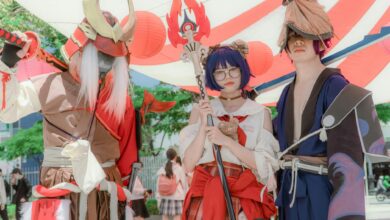 Three cosplayers dressed as fantasy characters pose together under vibrant canopy outdoors.
