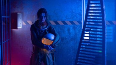 A female astronaut holding a helmet in a dramatic, futuristic setting with blue and red lighting.