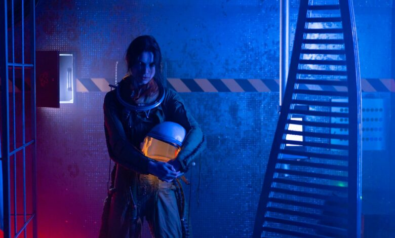 A female astronaut holding a helmet in a dramatic, futuristic setting with blue and red lighting.