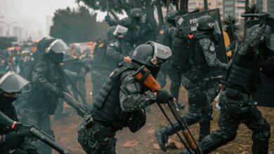 Riot police equipped with shields and rifles during a protest in an urban setting.