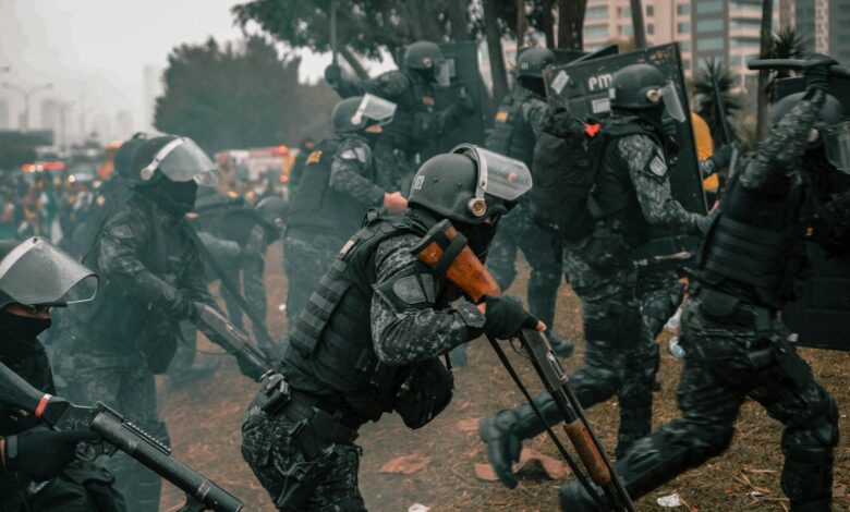 Riot police equipped with shields and rifles during a protest in an urban setting.