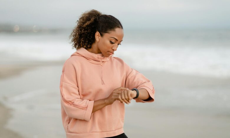 A woman in casual attire checks her smartwatch while standing on a sandy beach.