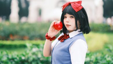 A young woman in cosplay holds a red apple in a vibrant, outdoor Vietnamese garden setting.