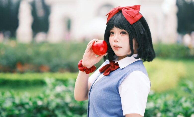 A young woman in cosplay holds a red apple in a vibrant, outdoor Vietnamese garden setting.