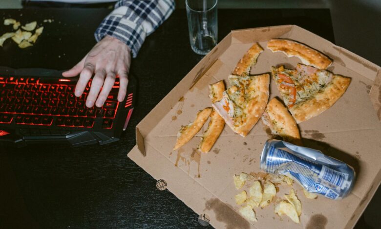 A gamer enjoys pizza and soda in a dimly lit room with a red-lit keyboard.