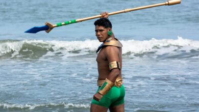 A muscular man in costume standing by the sea, holding a spear with waves in the background.