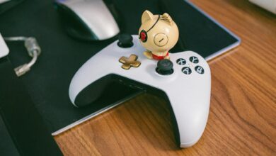 A charming cat figurine sits atop a gaming controller, beside a computer mouse, on a wooden desk.