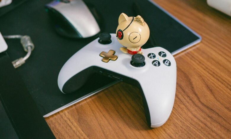A charming cat figurine sits atop a gaming controller, beside a computer mouse, on a wooden desk.