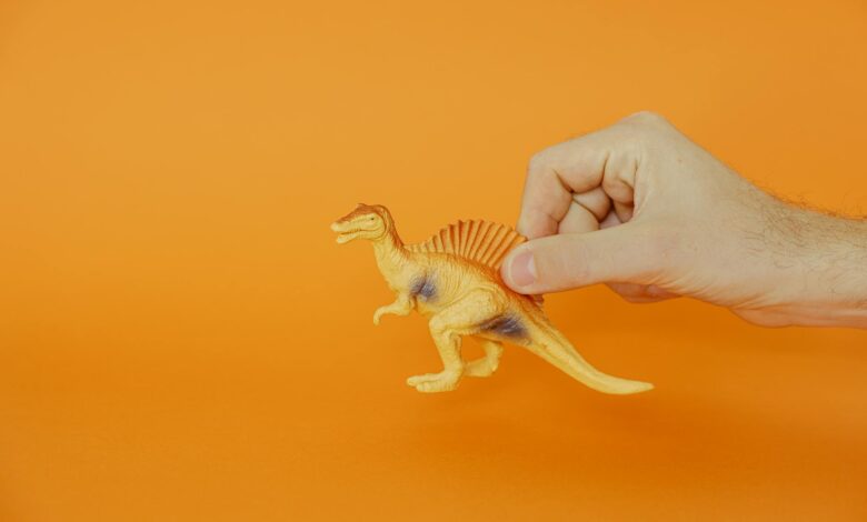 A hand holding a plastic dinosaur toy against a vibrant orange background, showcasing creativity and playfulness.