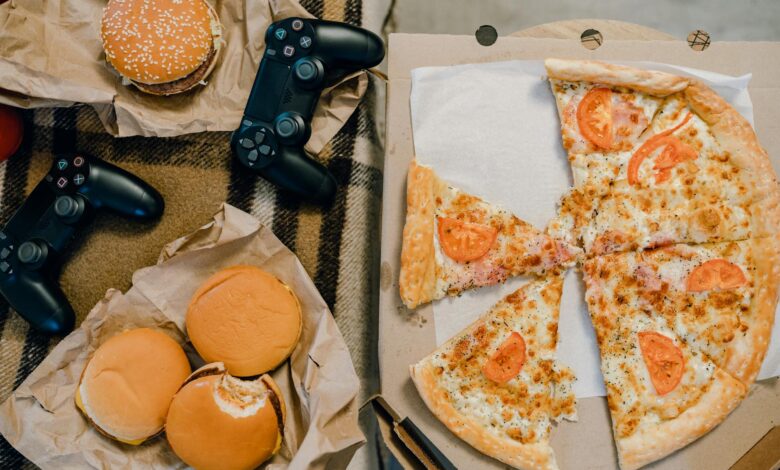 Aerial view of delicious pizza, burgers, and gaming controllers on a plaid blanket.