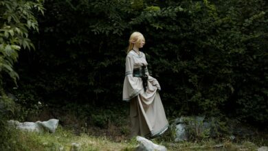 A cosplayer dressed as an elf in a fantasy costume stands in a lush, green forest.