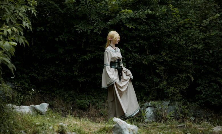A cosplayer dressed as an elf in a fantasy costume stands in a lush, green forest.