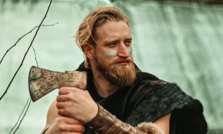 Bearded man dressed as a Viking with face paint, holding an axe outdoors.