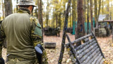A man wearing camouflage gear participates in an outdoor paintball game in a forested area.