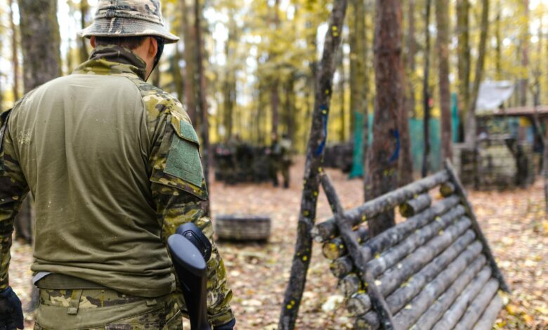 A man wearing camouflage gear participates in an outdoor paintball game in a forested area.
