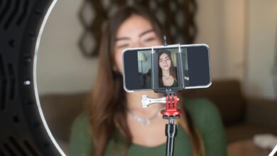 A young woman live streams from home using a smartphone and ring light setup.