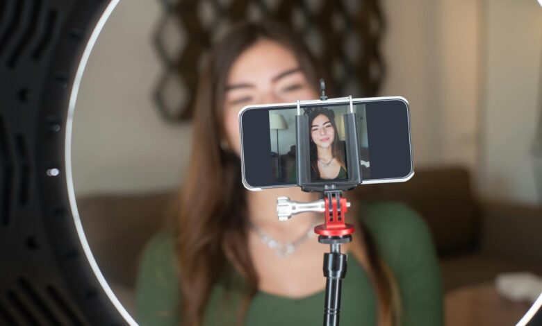 A young woman live streams from home using a smartphone and ring light setup.