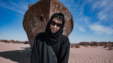 A man in dark clothing stands before a rusted ship in the sandy Aral Desert under a bright blue sky.