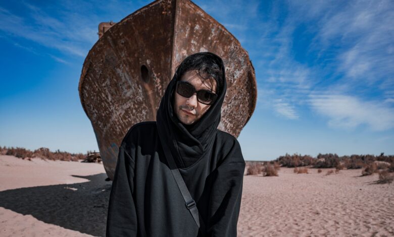 A man in dark clothing stands before a rusted ship in the sandy Aral Desert under a bright blue sky.