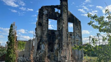 A desolated, burned-down house stands beneath a vibrant blue sky, symbolizing destruction and resilience.
