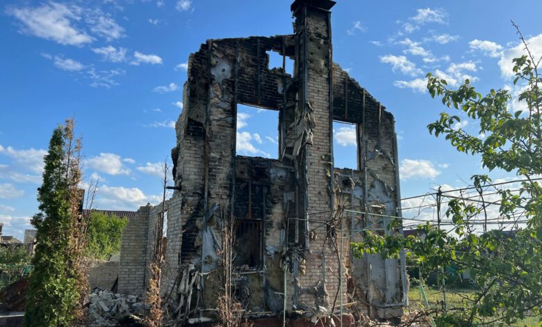 A desolated, burned-down house stands beneath a vibrant blue sky, symbolizing destruction and resilience.