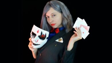 Side view of serious young female casino dealer in classy suit with bow tie holding face mask and cards in hands and looking at camera against black background