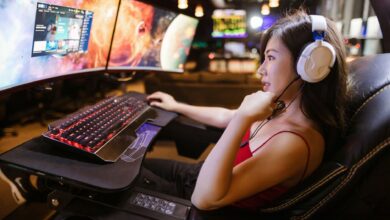 Young woman in red top gaming on dual monitors with white headphones in a modern setup.