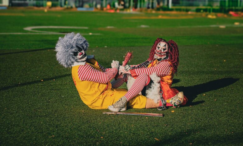 Two frightening clowns dressed in vibrant costumes sitting playfully on a sunny field.