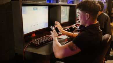 A young man is gaming using a smartphone and a PC setup with a mechanical keyboard indoors.