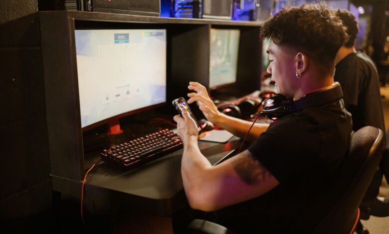 A young man is gaming using a smartphone and a PC setup with a mechanical keyboard indoors.
