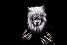 Striking portrait of an adult in a realistic werewolf costume with metallic claws against a black background.