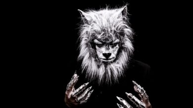 Striking portrait of an adult in a realistic werewolf costume with metallic claws against a black background.