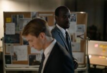Two detectives examining a corkboard filled with evidence in a dimly lit office.
