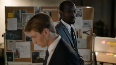 Two detectives examining a corkboard filled with evidence in a dimly lit office.