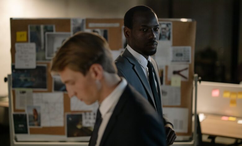 Two detectives examining a corkboard filled with evidence in a dimly lit office.