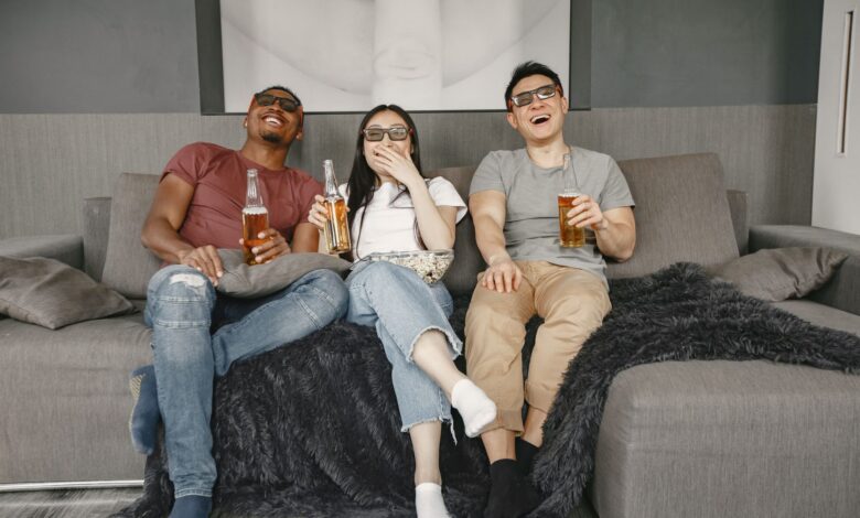Three friends relaxing on a sofa with drinks, watching a movie and laughing indoors.