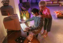 Two friends in a cozy room share a moment using a vintage desktop computer, illuminated softly.